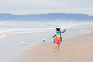 girl jumping on the beach with a rainbow cape