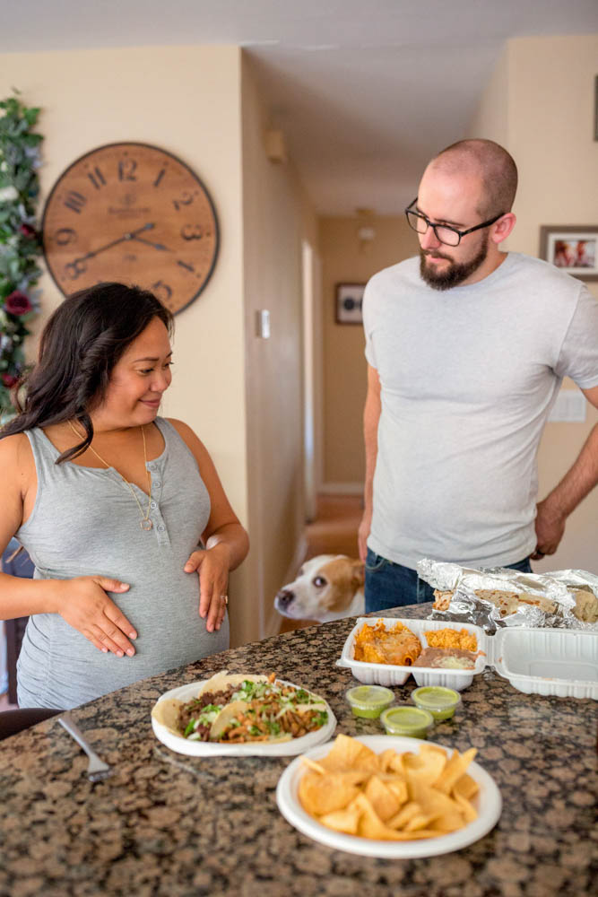 Pregnant mom rubbing belly, dad standing next to her, dog in background, mexican food on counter