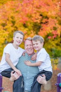 dad holding his two boys in his arms with fall leaves of orange and red in the background