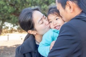 family photos in oakland with family of three with mom and dad kissing daughters cheeks