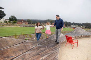little girl standing on wooden bench sculpture with her parents holding her hands at presidio tunnel tops