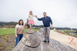 little girl standing on wooden bench sculpture with her parents holding her hands at presidio tunnel tops