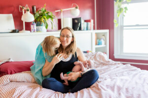 mom holding newborn and getting kiss on check from toddler