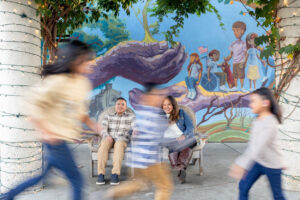 mom and dad sitting in chairs with a mural in the background at los altos downtown. the three kids are in slow motion walking and running in front of them